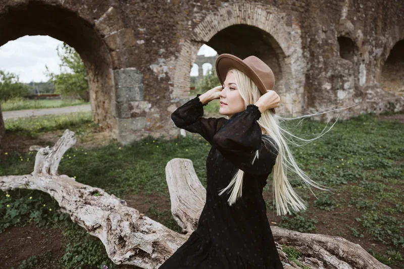 Modella con cappello e abito nero tra archi antichi e capelli mossi dal vento