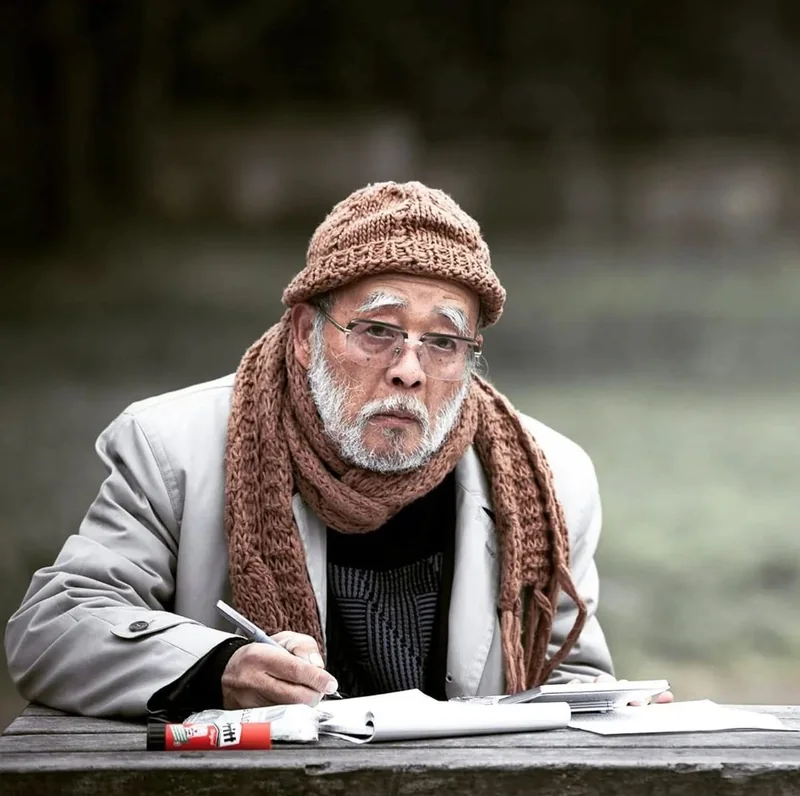 Uomo anziano con cappello e sciarpa, seduto a un tavolo all’aperto con un quaderno