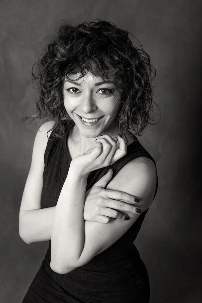 Ritratto in bianco e nero di una donna dai capelli ricci, sorridente con la mano sotto il mento