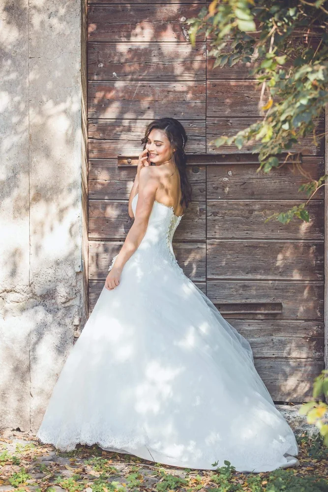 Sposa in abito bianco davanti a un portone di legno, tra ombre di foglie