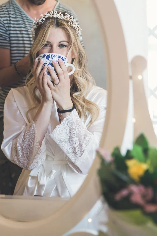 Sposa bionda in vestaglia bianca beve da una tazza decorata davanti allo specchio