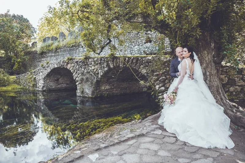 Coppia di sposi abbracciati sotto un albero accanto a un ponte di pietra e all’acqua