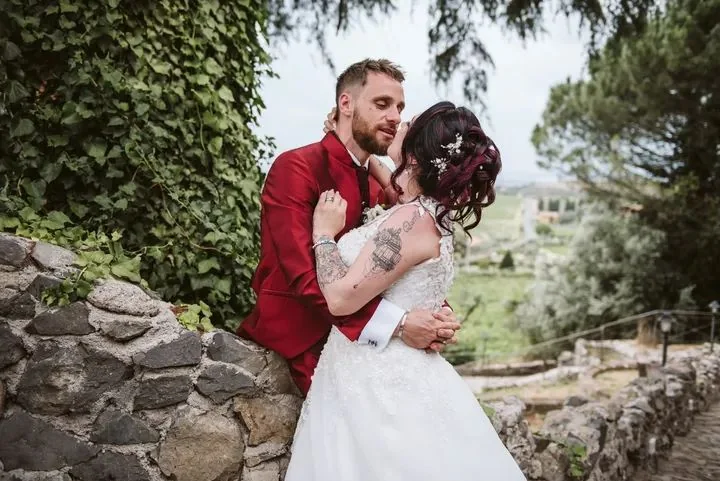 Sposo in giacca rossa e sposa tatuata si abbracciano accanto a un muretto di pietra